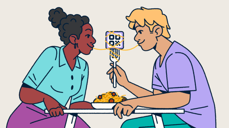 A couple sitting at a table sharing a bowl of spaghetti and meatballs. One partner is holding a fork with a QR Code on it and a piece of spaghetti wrapped around it. They each have one end of the noodle in their mouth.