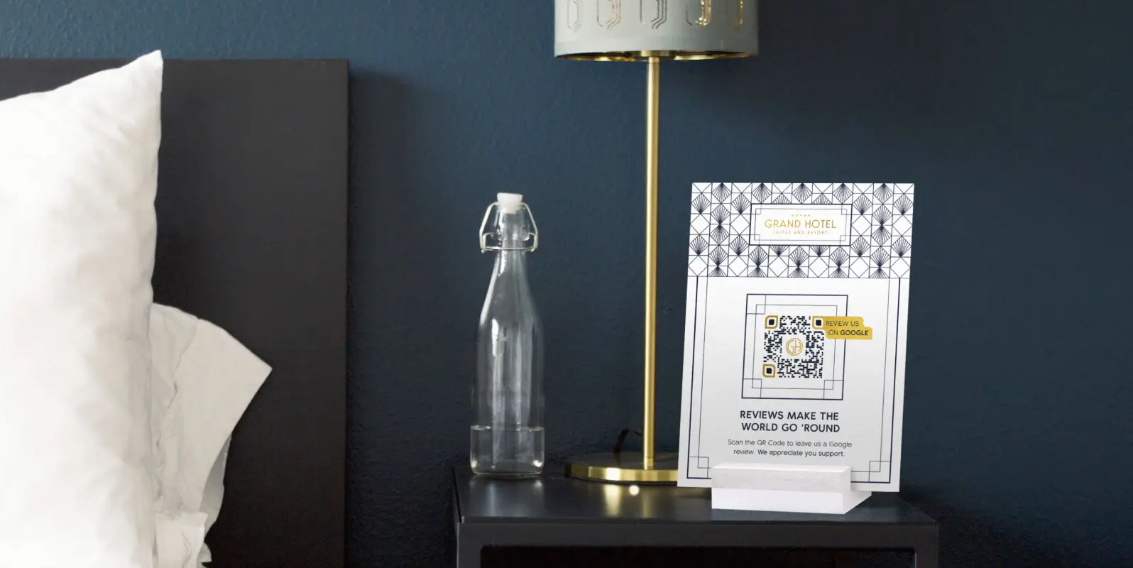 Hotel bedside table with a bottle of water, lamp, and sign asking for reviews on it.